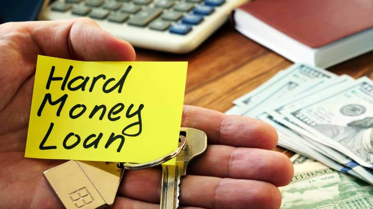 A hand holds a note that reads “Hard Money Loan”. Behind it, there is money on a table.