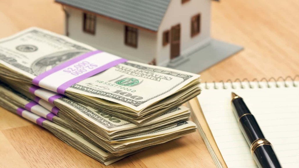 A stack of 100 dollar bills lay next to a miniature model house and a pen on top of paper.